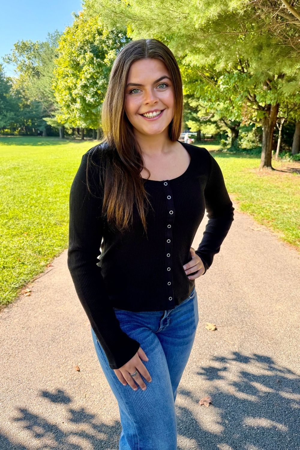 A woman smiling outdoors on a paved path in a park, wearing a black long-sleeve button-front top and blue jeans.