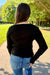 A woman viewed from behind wearing a black long-sleeve ribbed top and blue jeans, standing on a paved path in a park with green trees in the background.