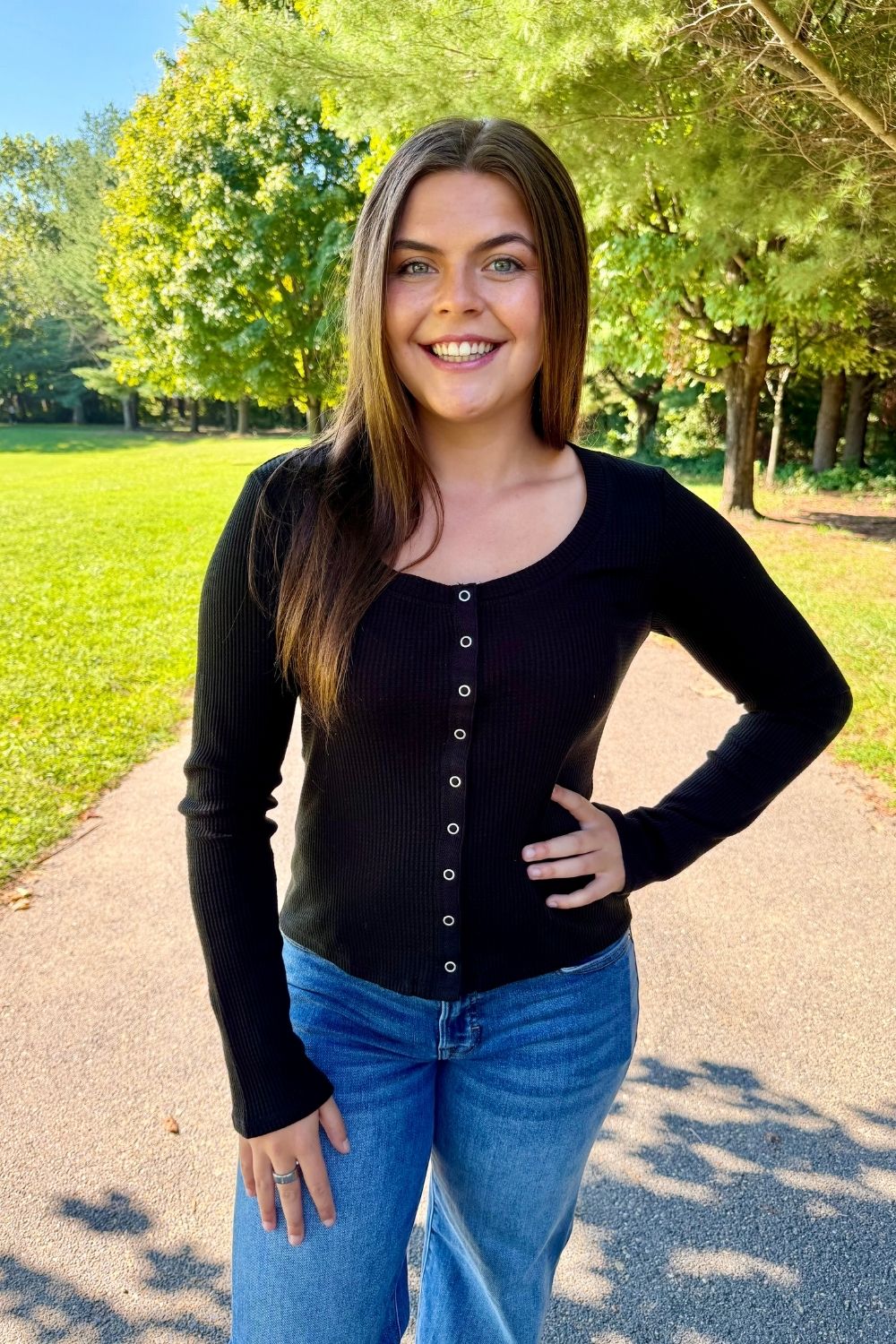 A smiling young woman with long brown hair stands outdoors on a paved path in a park, wearing a black ribbed long-sleeve top with a button-down front and blue jeans.