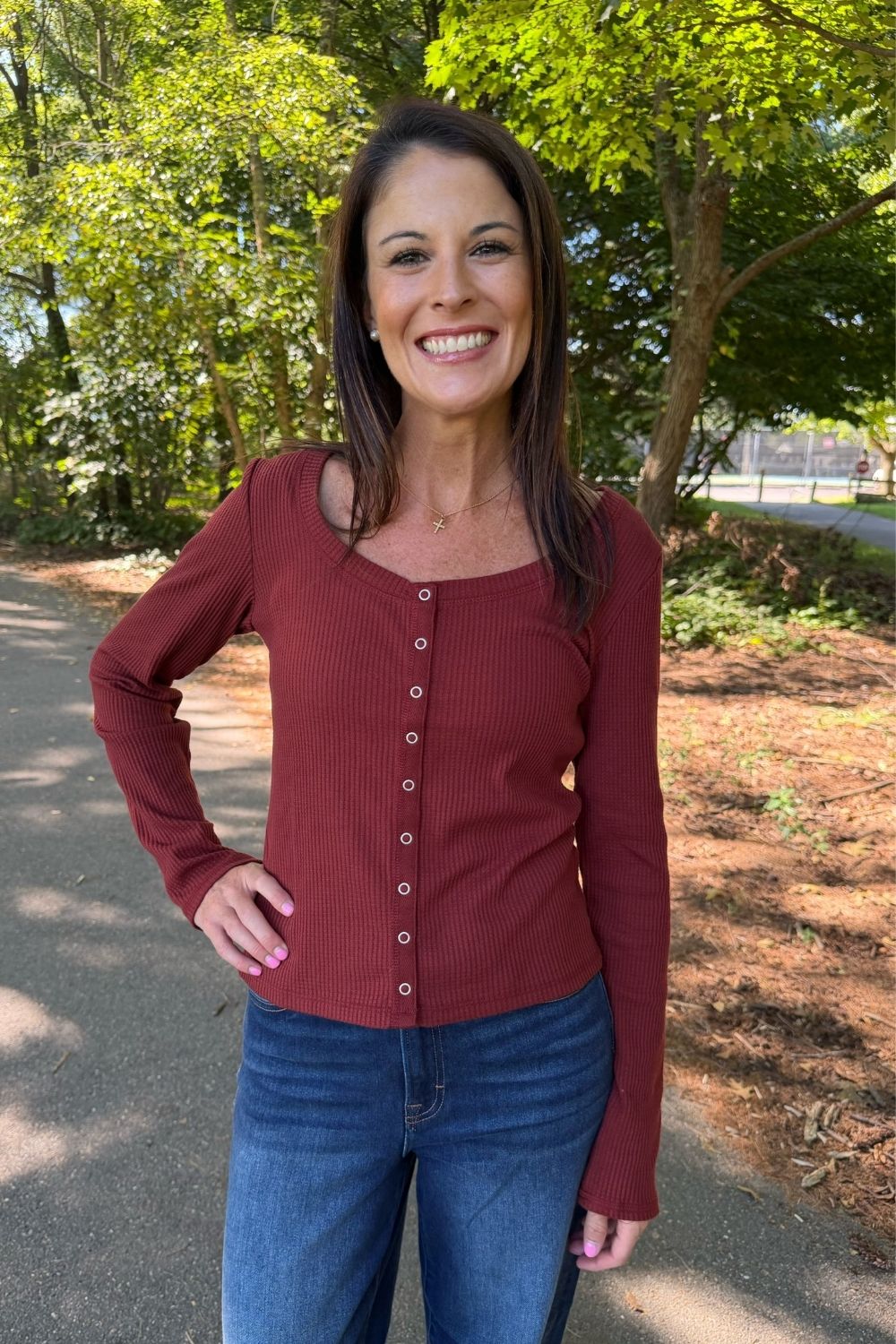 A woman smiling outdoors on a paved path wearing a rust-red ribbed long-sleeve button-front top and blue jeans, with trees in the background.