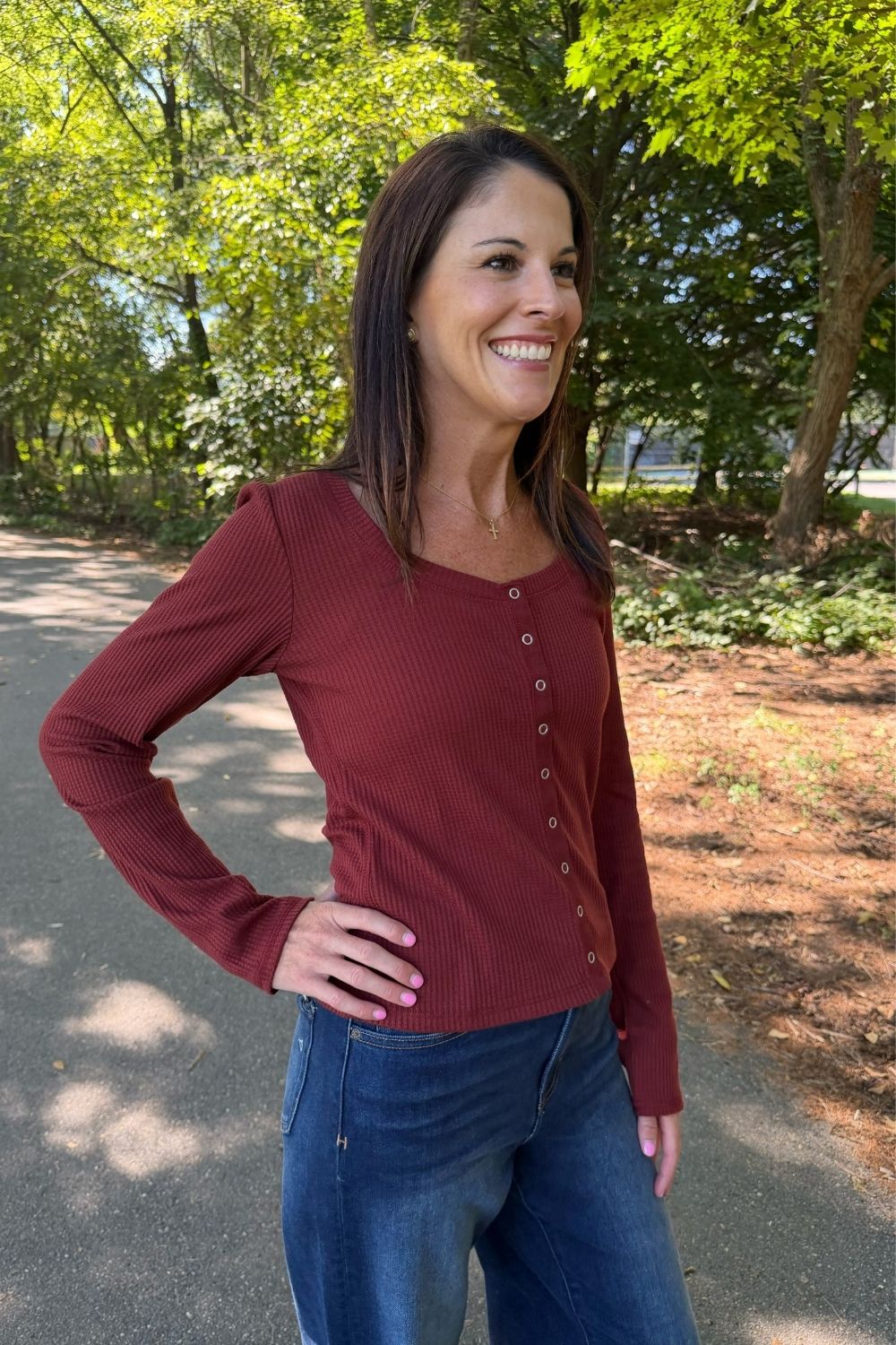 A woman wearing a long-sleeve ribbed button-front top in burgundy and blue jeans, standing outdoors on a paved path with trees in the background.