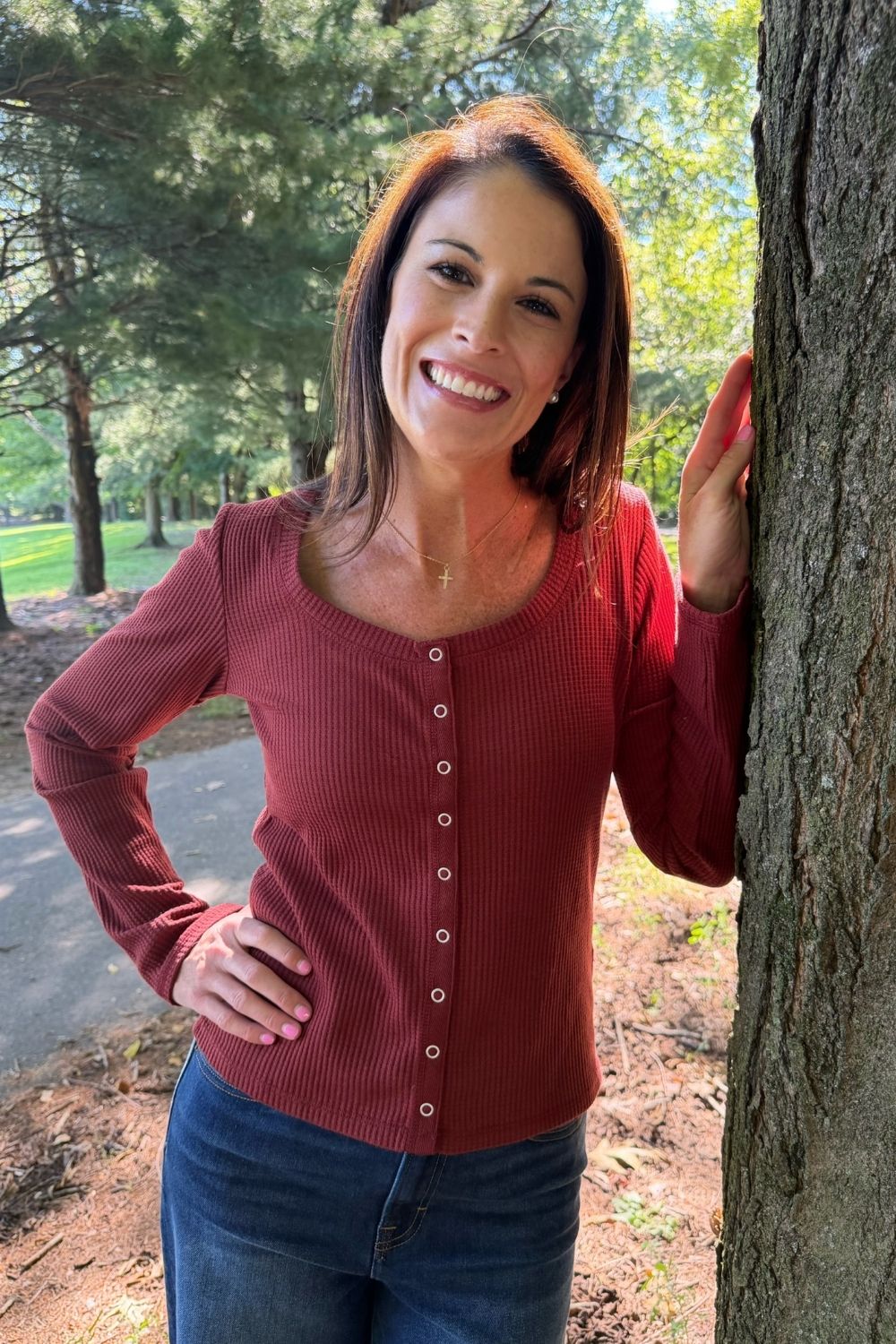 A woman smiling and posing outdoors next to a tree, wearing a long-sleeve ribbed button-front top in a muted brick red color and blue jeans.