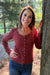 A woman smiling and posing outdoors next to a tree, wearing a long-sleeve ribbed button-front top in a muted brick red color and blue jeans.