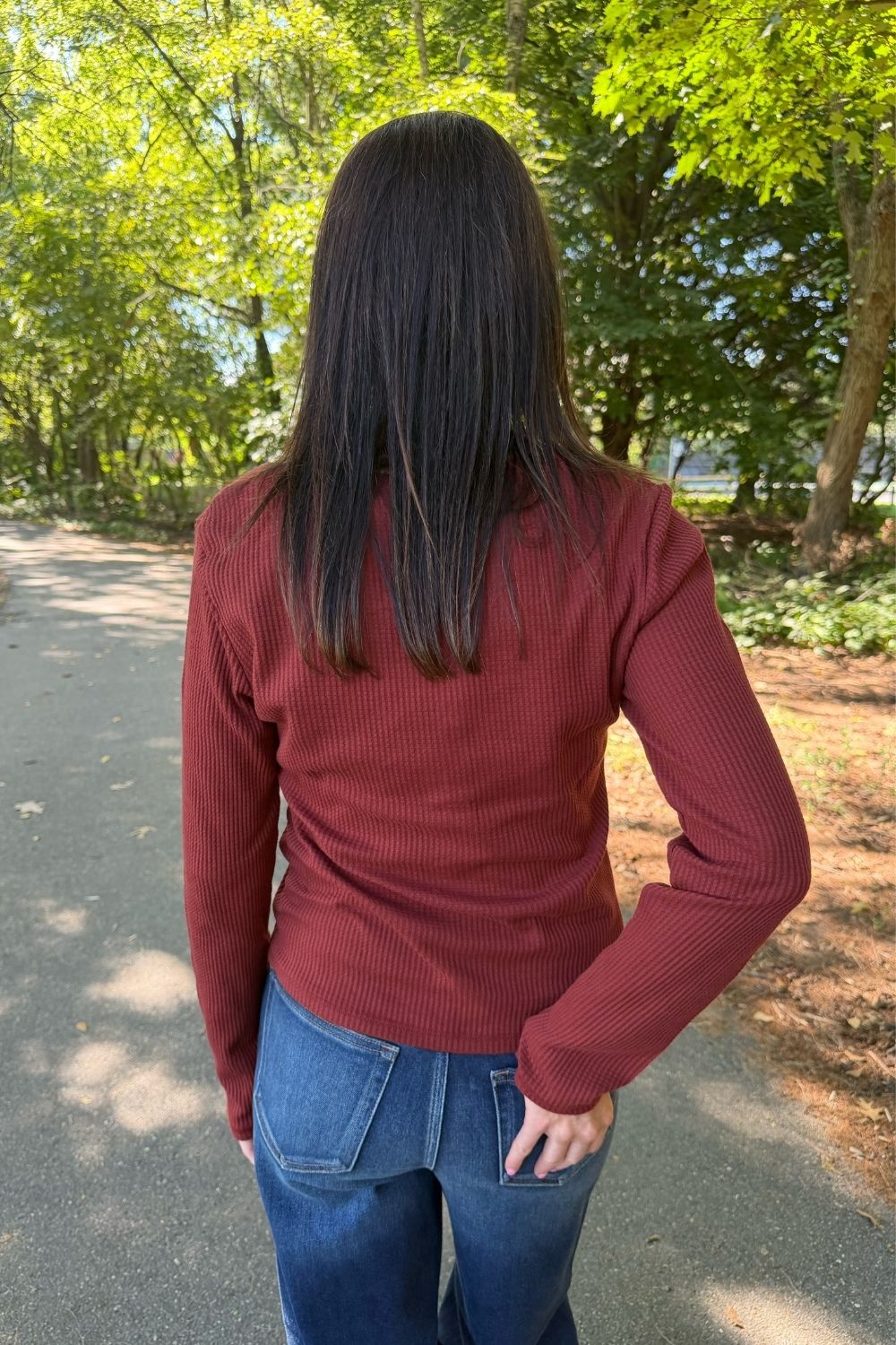 A woman viewed from behind wearing a rust-red ribbed long-sleeve top and blue jeans, standing on a paved path outdoors with trees in the background.