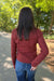 A woman viewed from behind wearing a rust-red ribbed long-sleeve top and blue jeans, standing on a paved path outdoors with trees in the background.