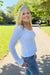 A smiling blonde woman wearing a white ribbed long-sleeve top and blue jeans stands on a paved path in a sunny park with green grass and trees in the background.