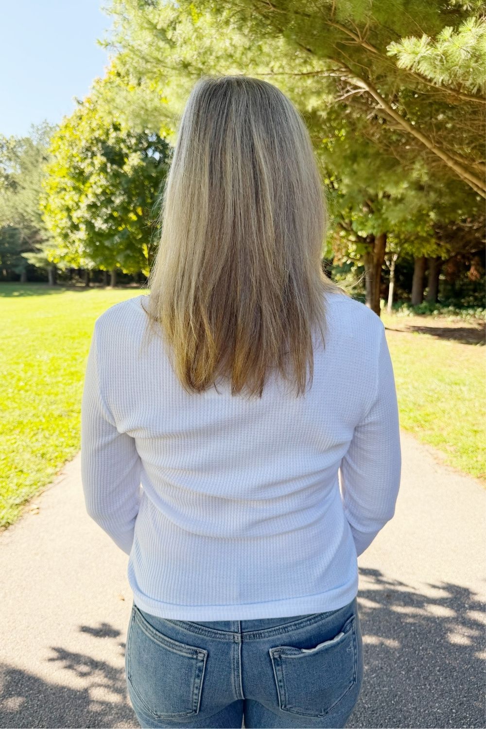 A woman viewed from behind wearing a white ribbed long-sleeve top and blue jeans, standing on a paved path in a sunny park with green grass and trees in the background.