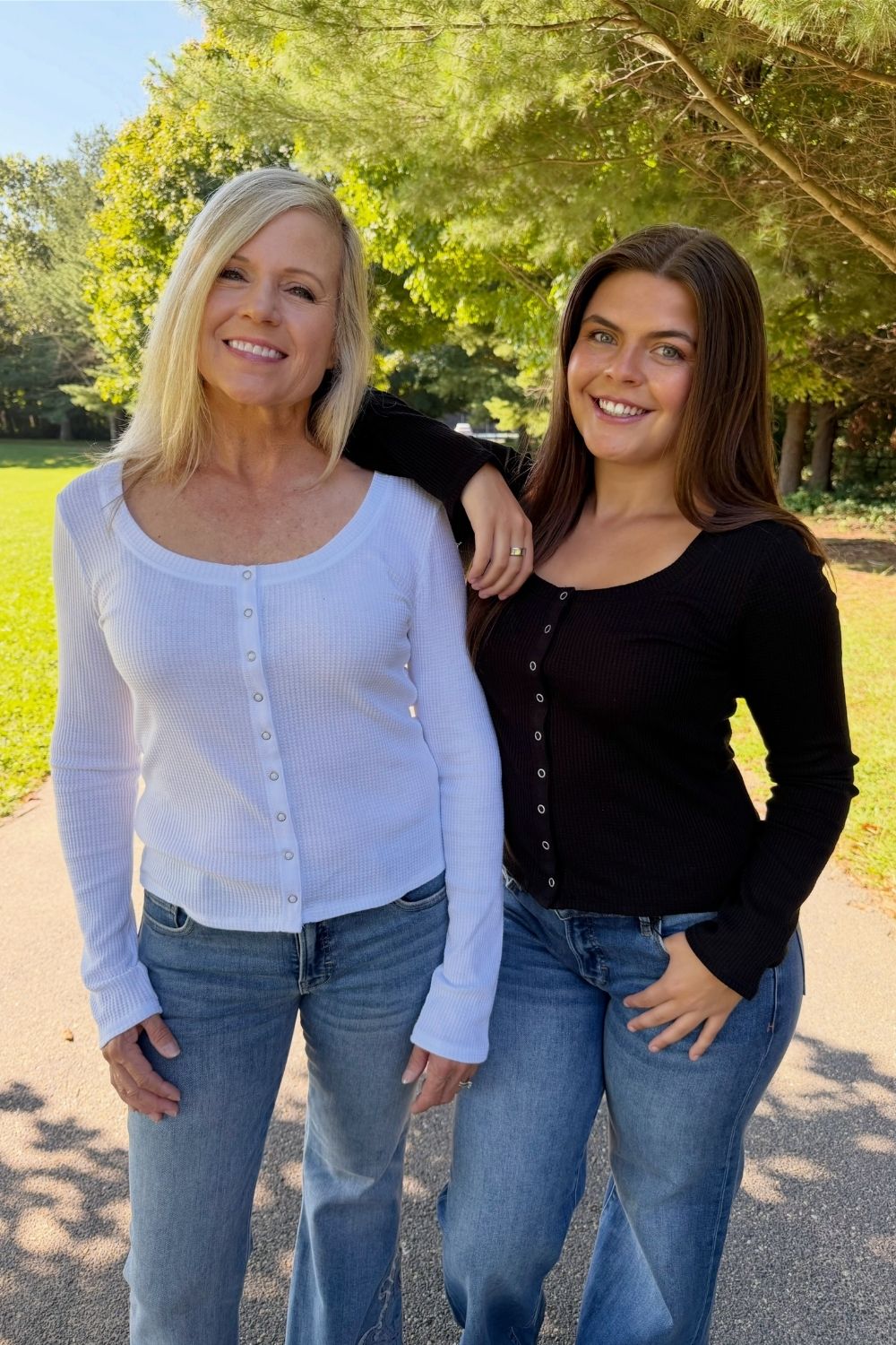 Two women standing outdoors on a paved path with trees and grass in the background. The woman on the left has blonde hair and wears a white ribbed long-sleeve top with blue jeans.