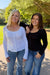 Two women standing outdoors on a paved path with trees and grass in the background. The woman on the left has blonde hair and wears a white ribbed long-sleeve top with blue jeans.
