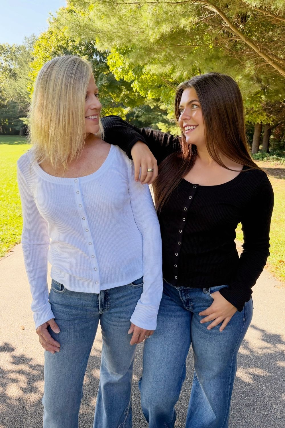 Two women standing outdoors on a paved path in a park, smiling at each other. The woman on the left wears a white long-sleeve button-front top with blue jeans, and the woman on the right wears a black long-sleeve button-front top with blue jeans.