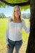 A smiling blonde woman wearing a white ribbed long-sleeve button-front top and blue jeans, standing outdoors next to a tree in a grassy park.