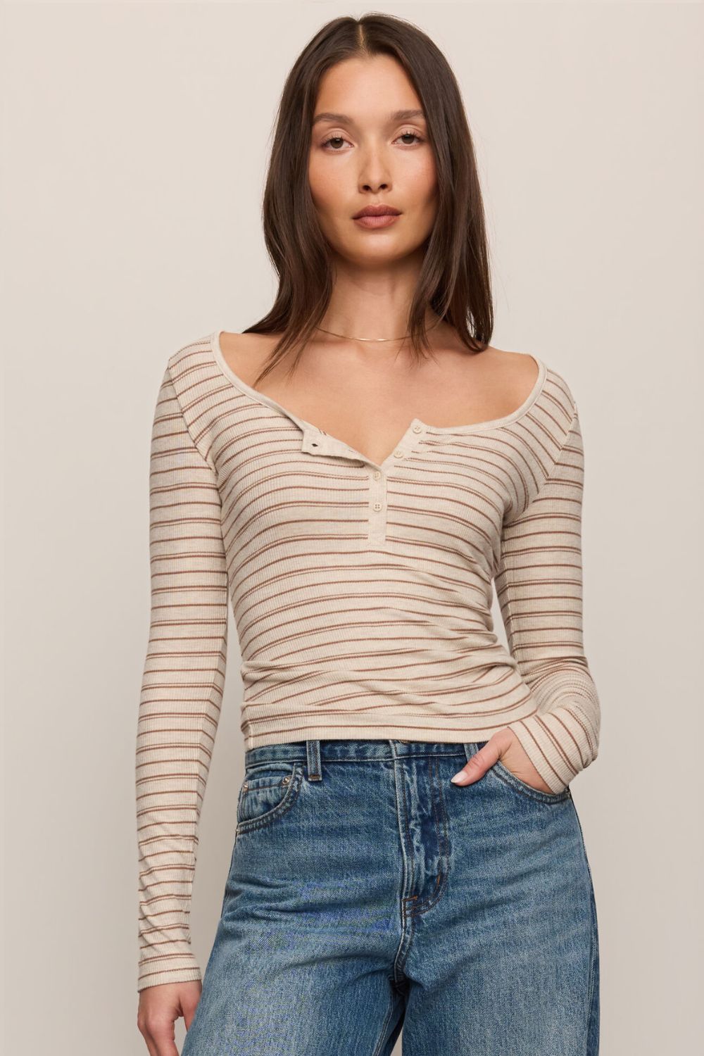 A woman modeling a beige and brown horizontally striped long-sleeve henley top with a wide scoop neckline, paired with blue jeans, against a solid light beige background.