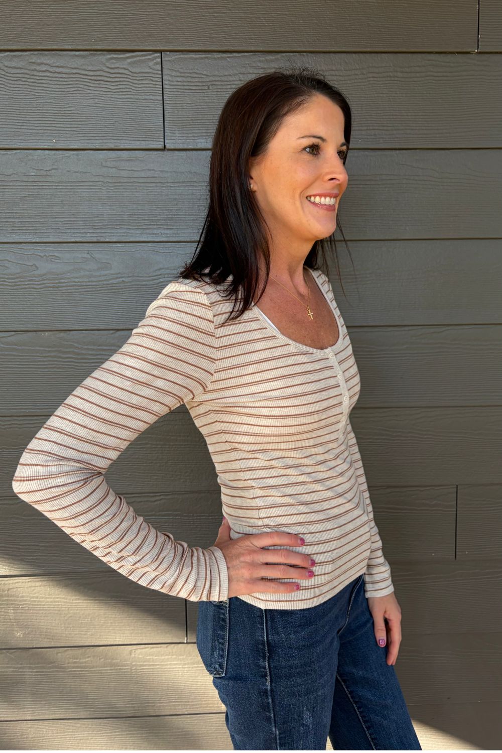 A woman wearing a beige long-sleeve top with thin brown horizontal stripes and blue jeans, standing against a gray wood-paneled wall.