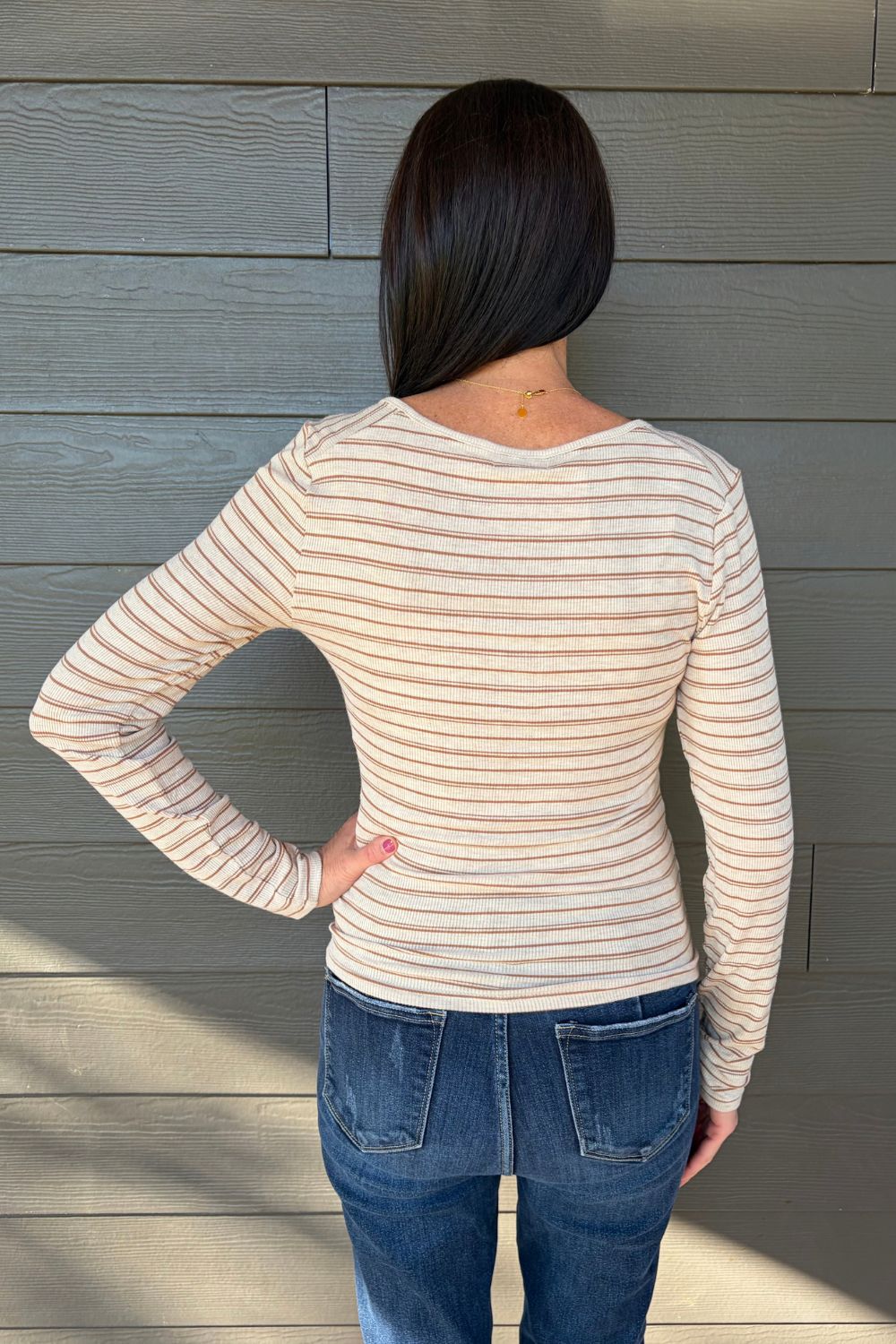 A woman viewed from behind wearing a beige long-sleeve top with thin brown horizontal stripes and blue jeans, standing against a gray wood-paneled wall.