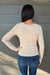 A woman viewed from behind wearing a beige long-sleeve top with thin brown horizontal stripes and blue jeans, standing against a gray wood-paneled wall.