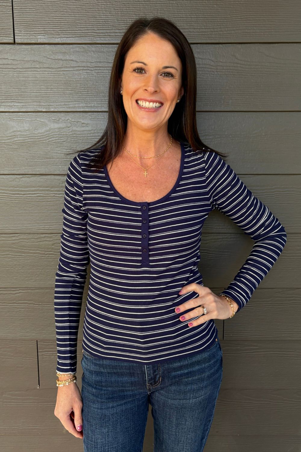 A woman wearing a navy blue and white striped long-sleeve V-neck top with blue jeans, posing against a gray wood-paneled wall.