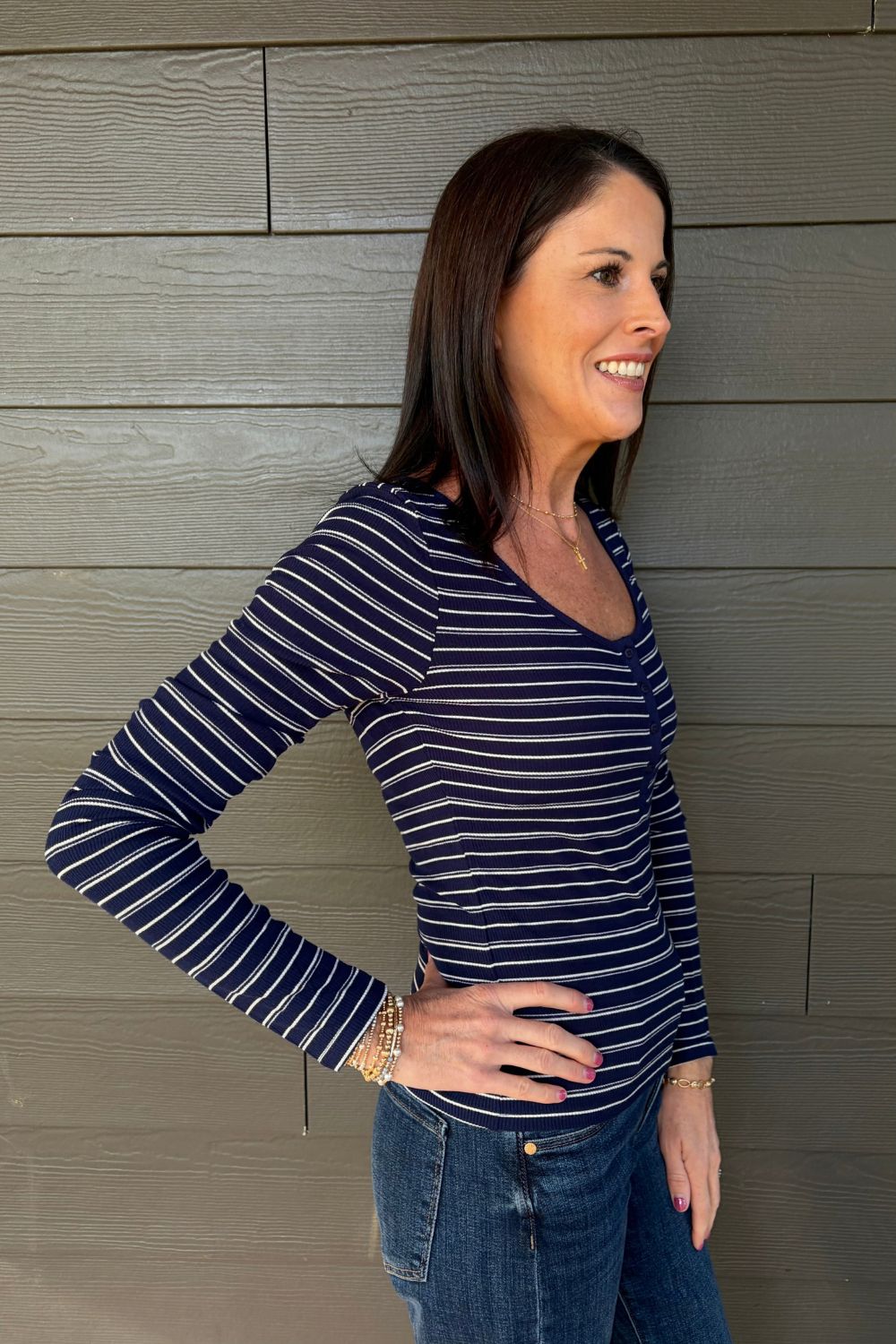 A woman wearing a navy blue and white striped long-sleeve V-neck top with blue jeans, posing against a gray wood-paneled wall.