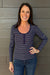 A woman wearing a navy blue and white striped long-sleeve V-neck top with blue jeans, posing against a gray wood-paneled wall.