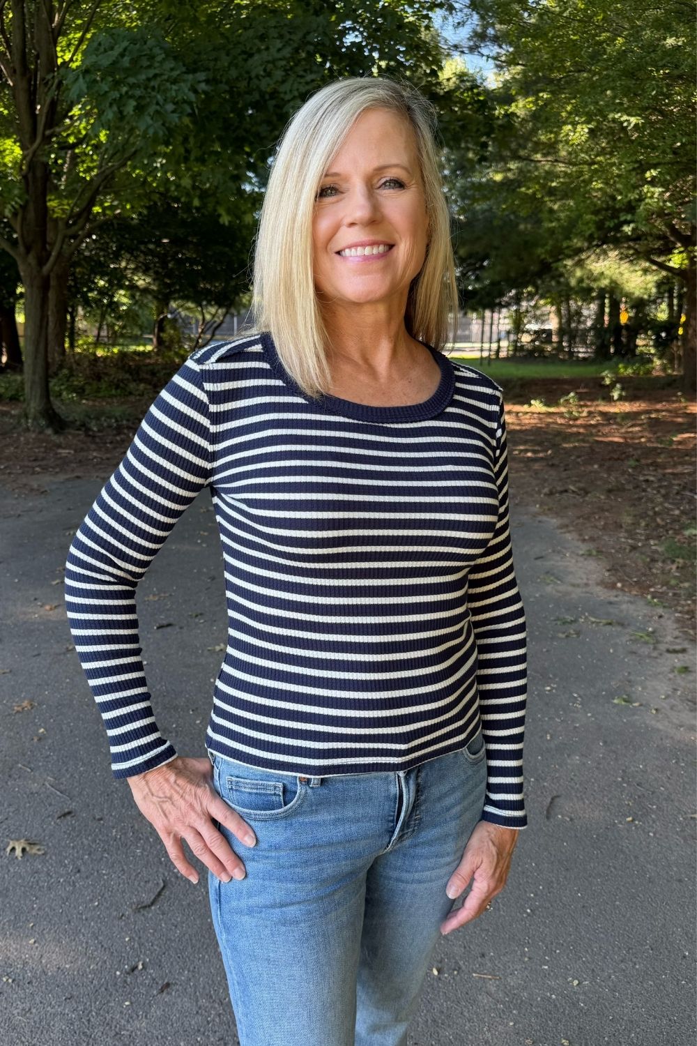 A smiling blonde woman wearing a navy and white horizontally striped long-sleeve top with blue jeans, standing outdoors on a paved path with trees in the background.