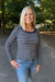A smiling blonde woman wearing a navy and white horizontally striped long-sleeve top with blue jeans, standing outdoors on a paved path with trees in the background.