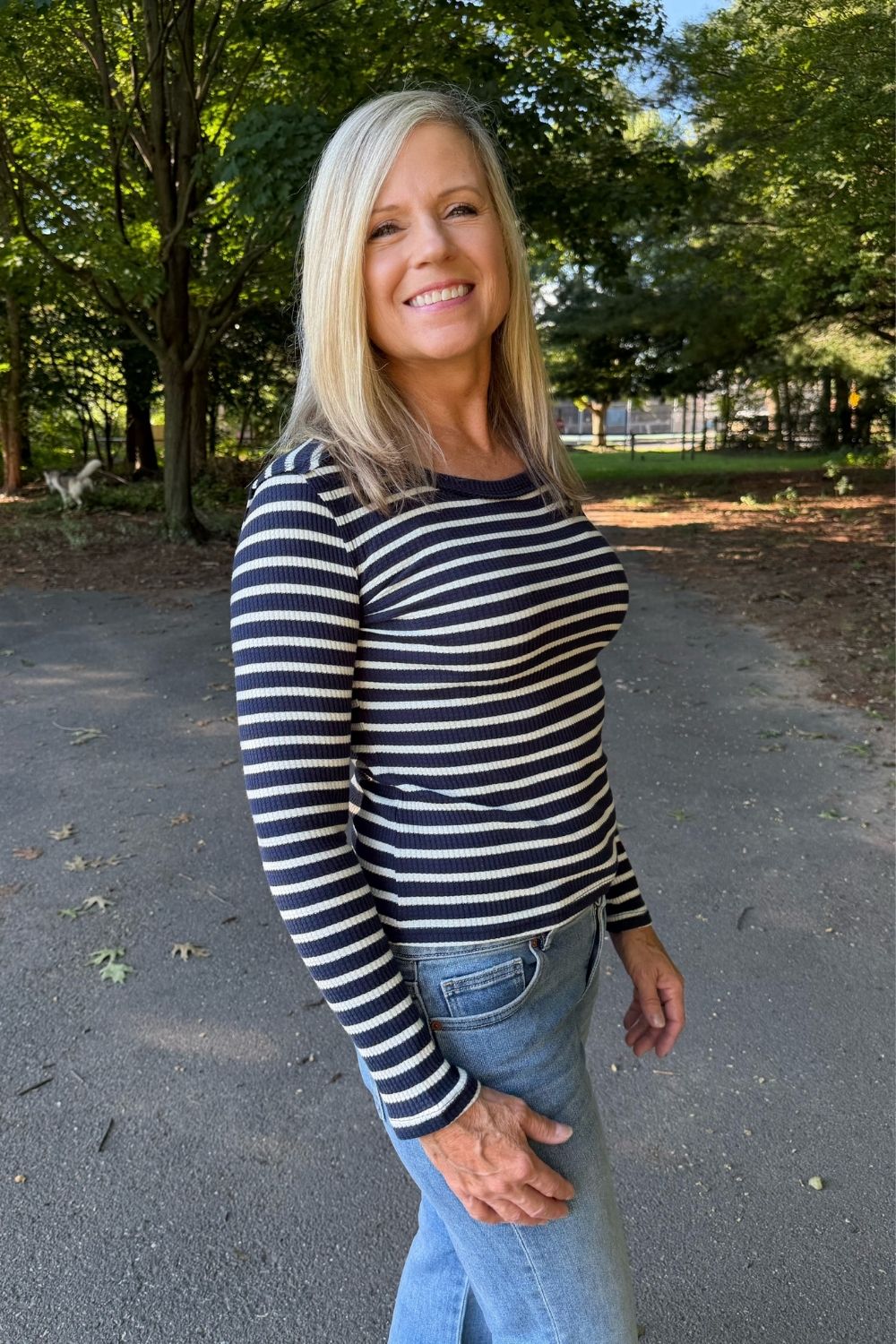 A smiling blonde woman wearing a navy and white horizontally striped long-sleeve top and light blue jeans, standing outdoors on a paved path with trees in the background.