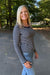 A smiling blonde woman wearing a navy and white horizontally striped long-sleeve top and light blue jeans, standing outdoors on a paved path with trees in the background.