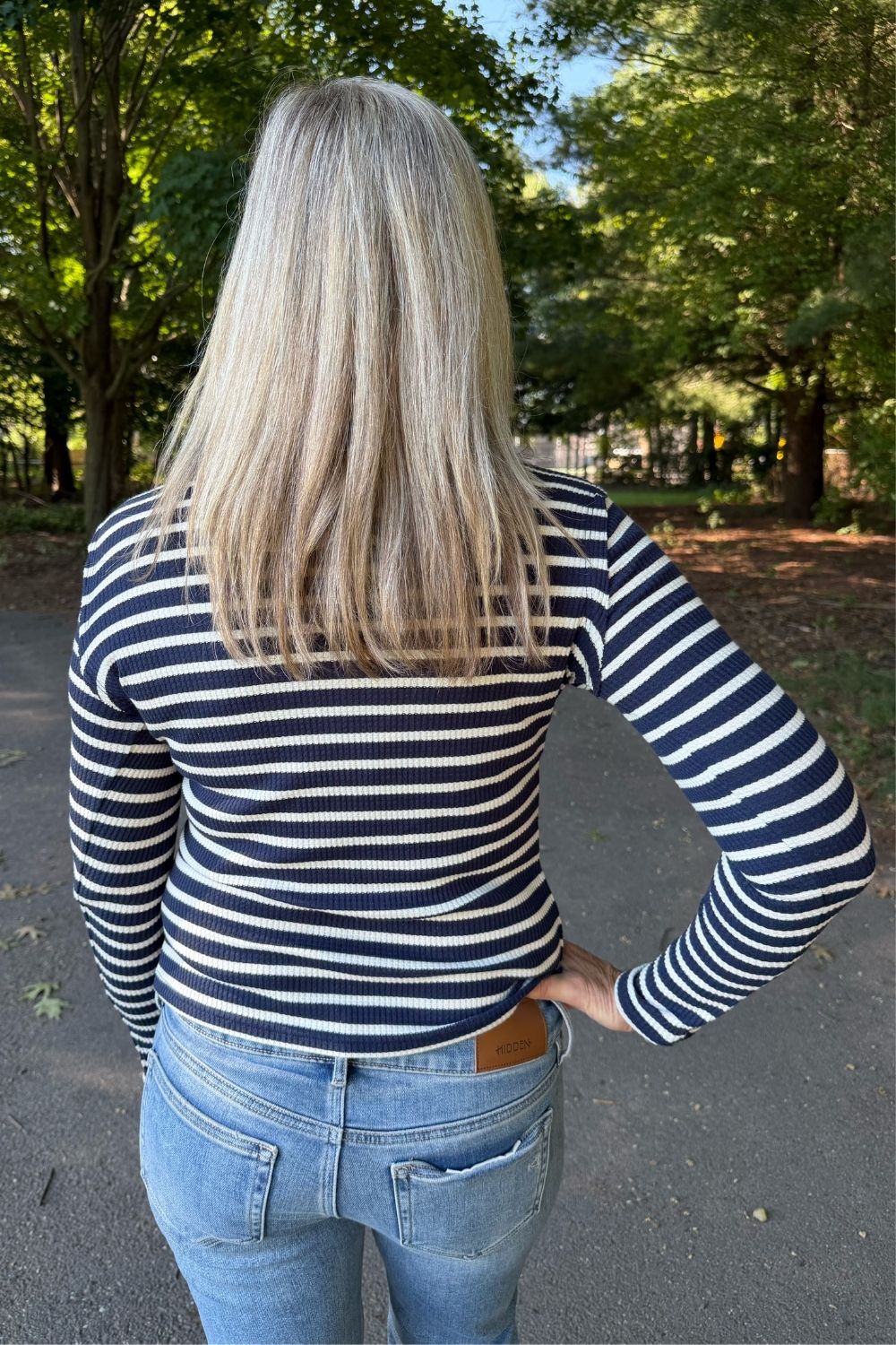 A woman viewed from behind wearing a navy and white horizontally striped long-sleeve top and light blue jeans, standing on an outdoor paved path with trees in the background.