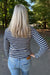 A woman viewed from behind wearing a navy and white horizontally striped long-sleeve top and light blue jeans, standing on an outdoor paved path with trees in the background.