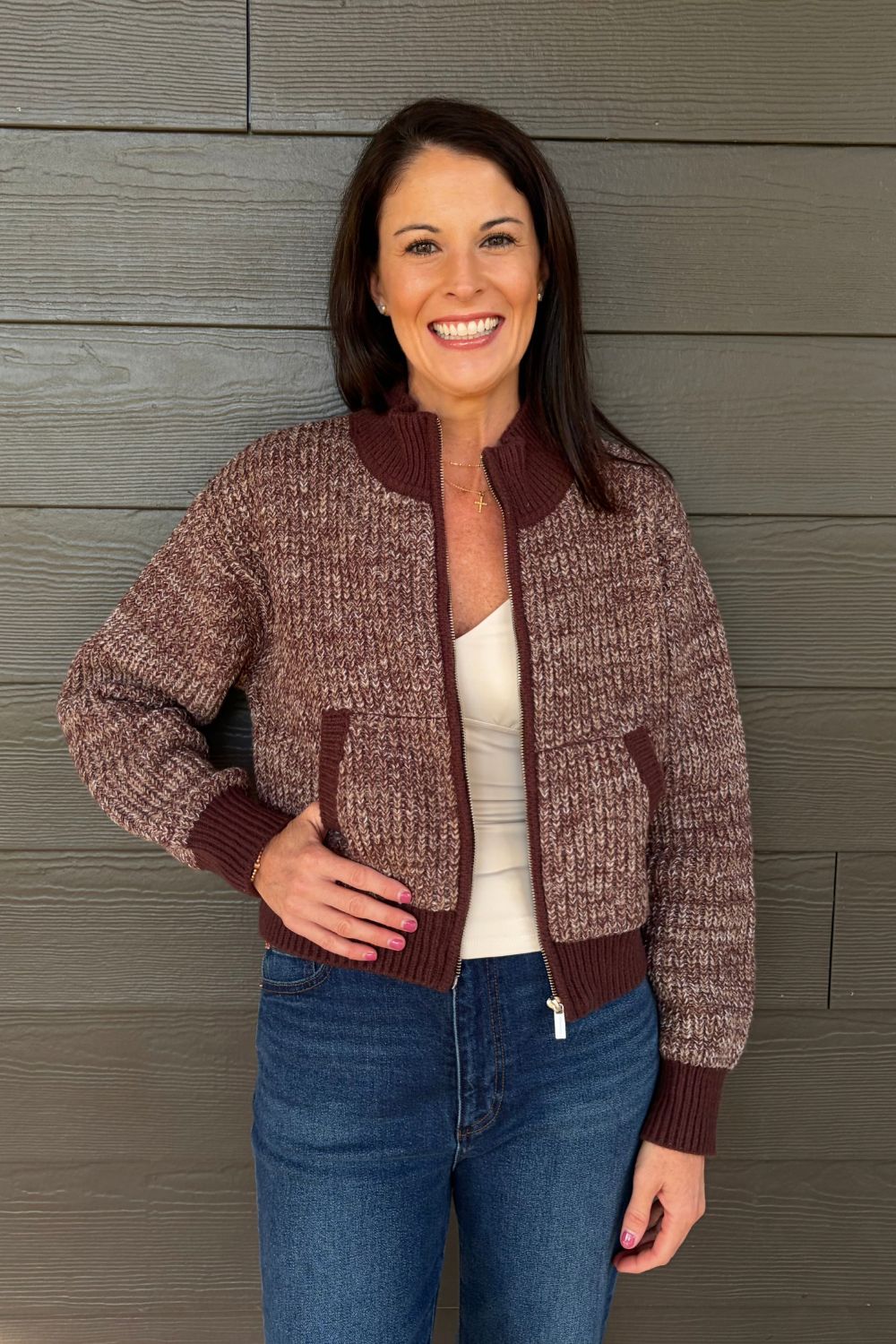 A woman wearing a marled brown and burgundy knit zip-up bomber jacket over a white top and blue jeans, standing against a gray wood-paneled wall.