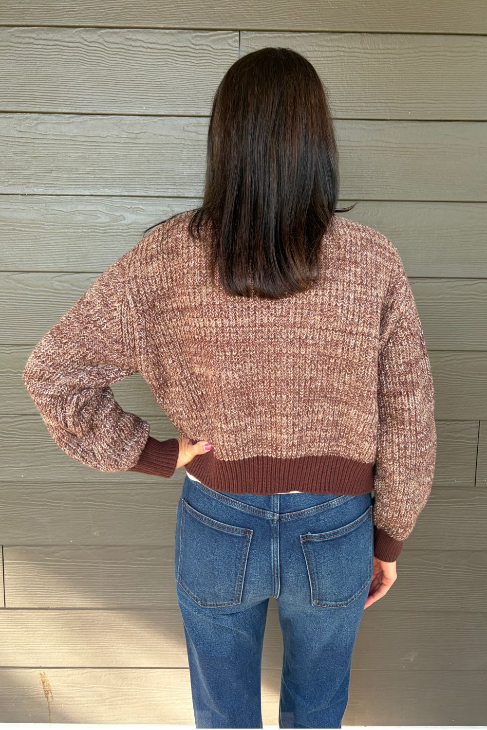 A person shown from the back wearing a brown and beige marled knit sweater with dark brown ribbed cuffs and hem, paired with blue jeans, standing against a gray wood-paneled wall.