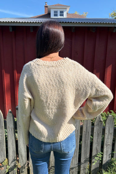 A woman wearing a cream-colored, textured knit sweater and blue jeans, posing outdoors in front of a red wooden fence and a weathered wooden picket fence.