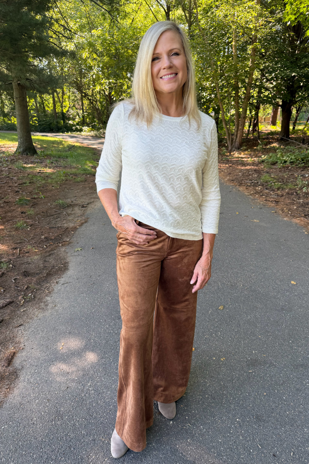 A woman standing on an outdoor paved path wearing a cream textured knit sweater and brown wide-leg pants.