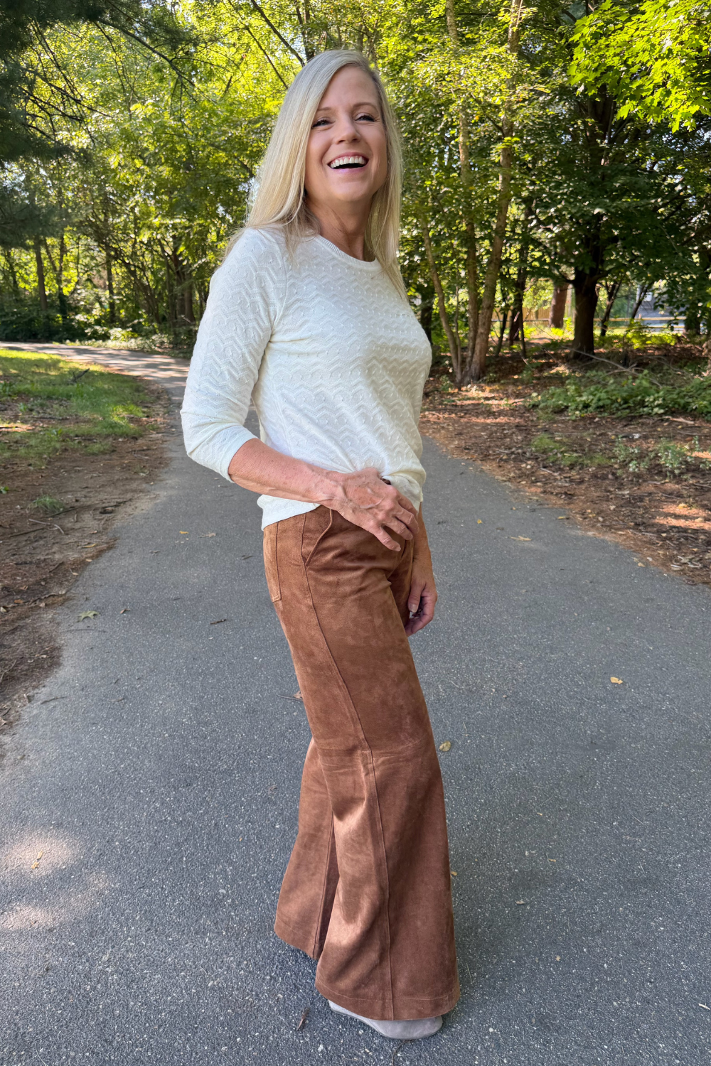 A woman stands on an outdoor paved path surrounded by trees, wearing a cream knit long-sleeve top and brown suede-like flared pants.