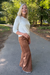 A woman stands on an outdoor paved path surrounded by trees, wearing a cream knit long-sleeve top and brown suede-like flared pants.