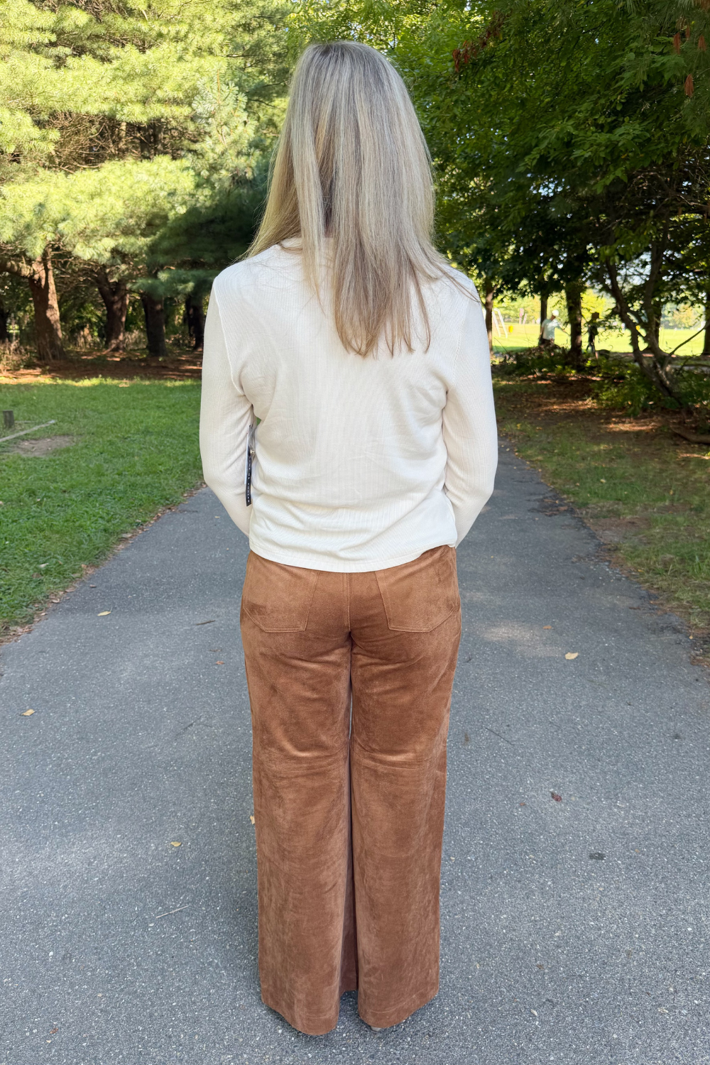 A woman viewed from behind wearing a cream ribbed long-sleeve top and brown wide-leg pants, standing on a paved path in a park with trees and grass in the background.