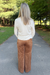 A woman viewed from behind wearing a cream ribbed long-sleeve top and brown wide-leg pants, standing on a paved path in a park with trees and grass in the background.