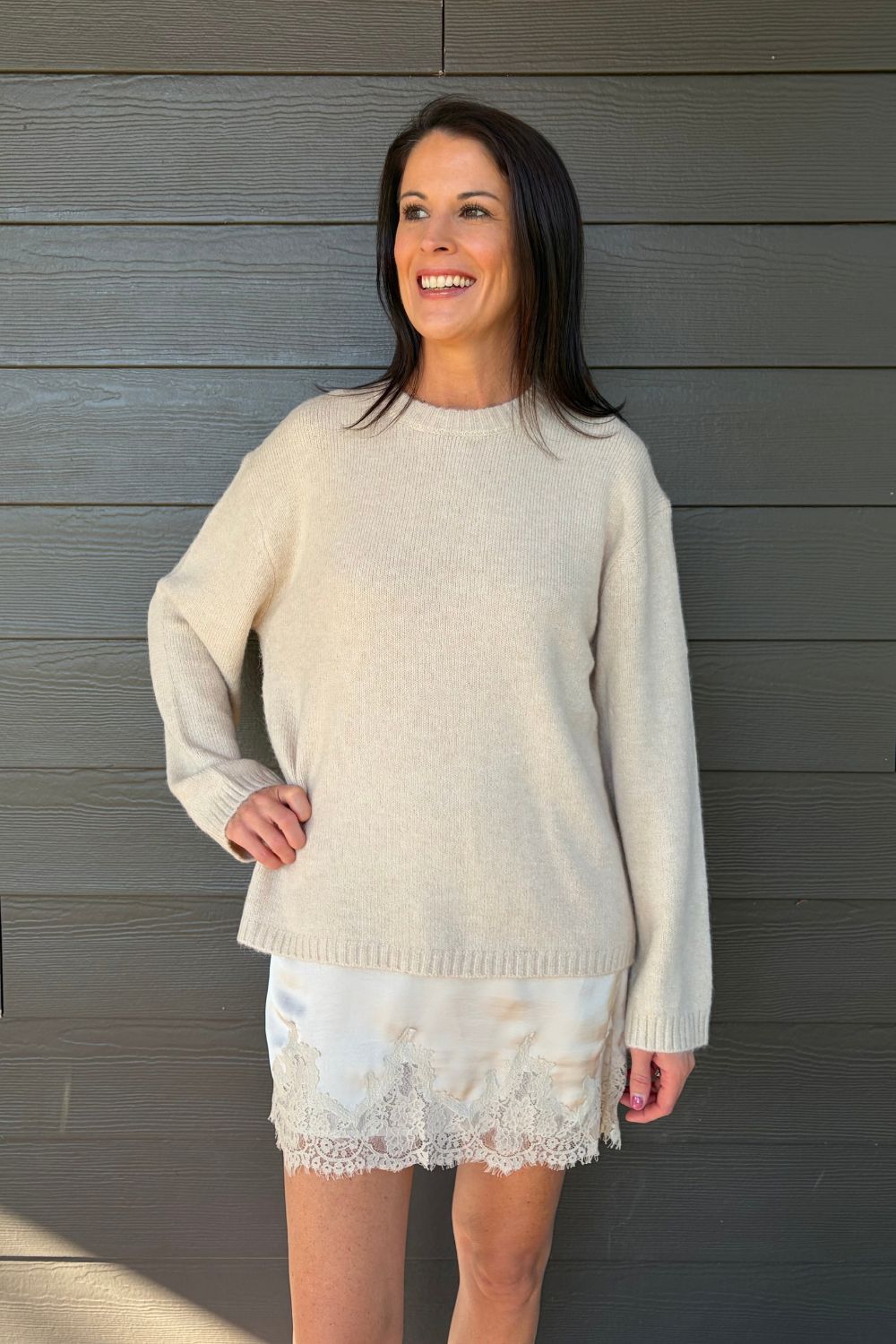 A woman wearing a cream-colored knit sweater and matching lace-trim shorts, posing against a dark gray wood-paneled wall.