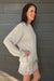 A woman wearing a cream-colored knit sweater and matching lace-trim shorts, posing against a dark gray wood-paneled wall.