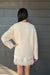 A person shown from the back wearing an oversized cream knit sweater and a short cream lace-trim dress or skirt, standing against a dark gray wood plank wall.