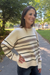 A woman wearing a cream and taupe striped knit sweater with black stripes, standing on an outdoor paved path with trees in the background.