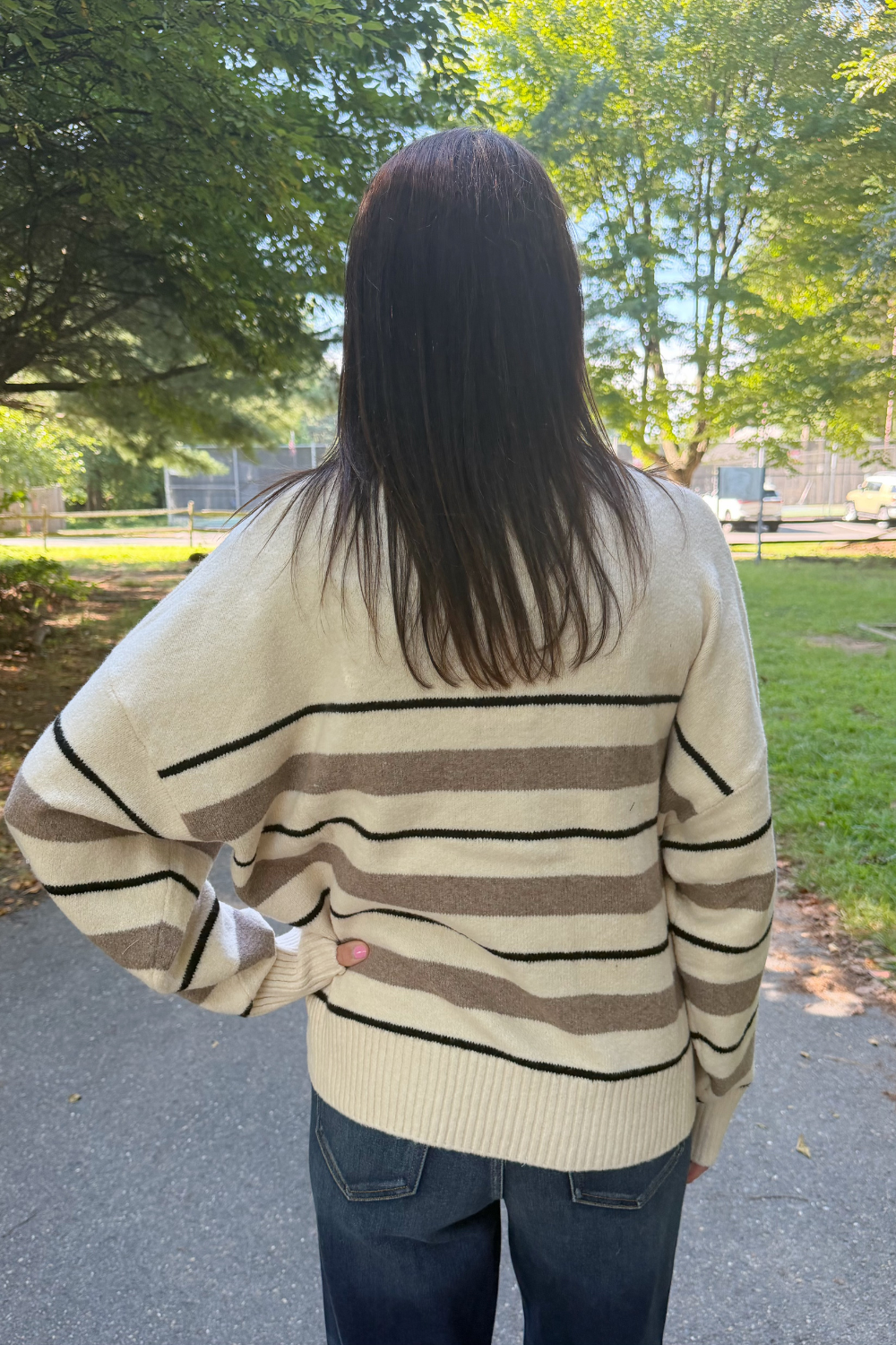 A person viewed from behind wearing a cream-colored knit sweater with brown and black horizontal stripes, paired with dark blue jeans, standing outdoors on a paved path with trees and grass in the background.