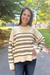 A woman smiling outdoors on a paved path wearing a cream-colored knit sweater with horizontal brown and black stripes, paired with dark blue jeans.