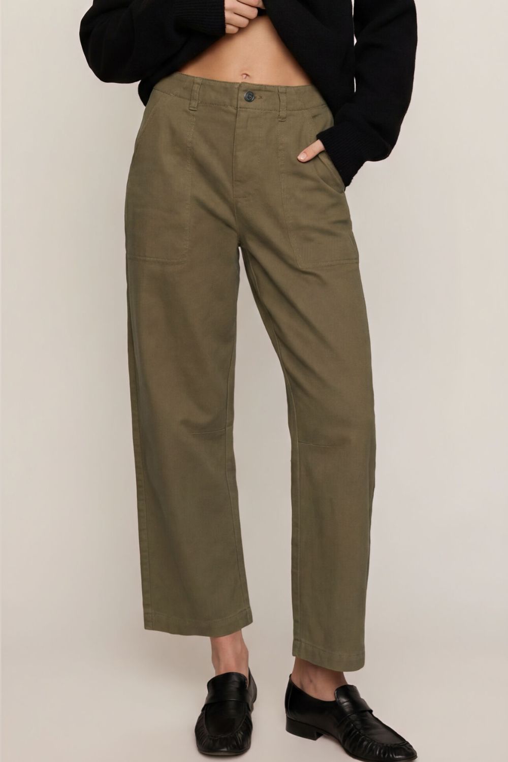 A person wearing olive green cropped trousers with a black cropped cardigan and black loafers, standing against a plain light beige studio background.