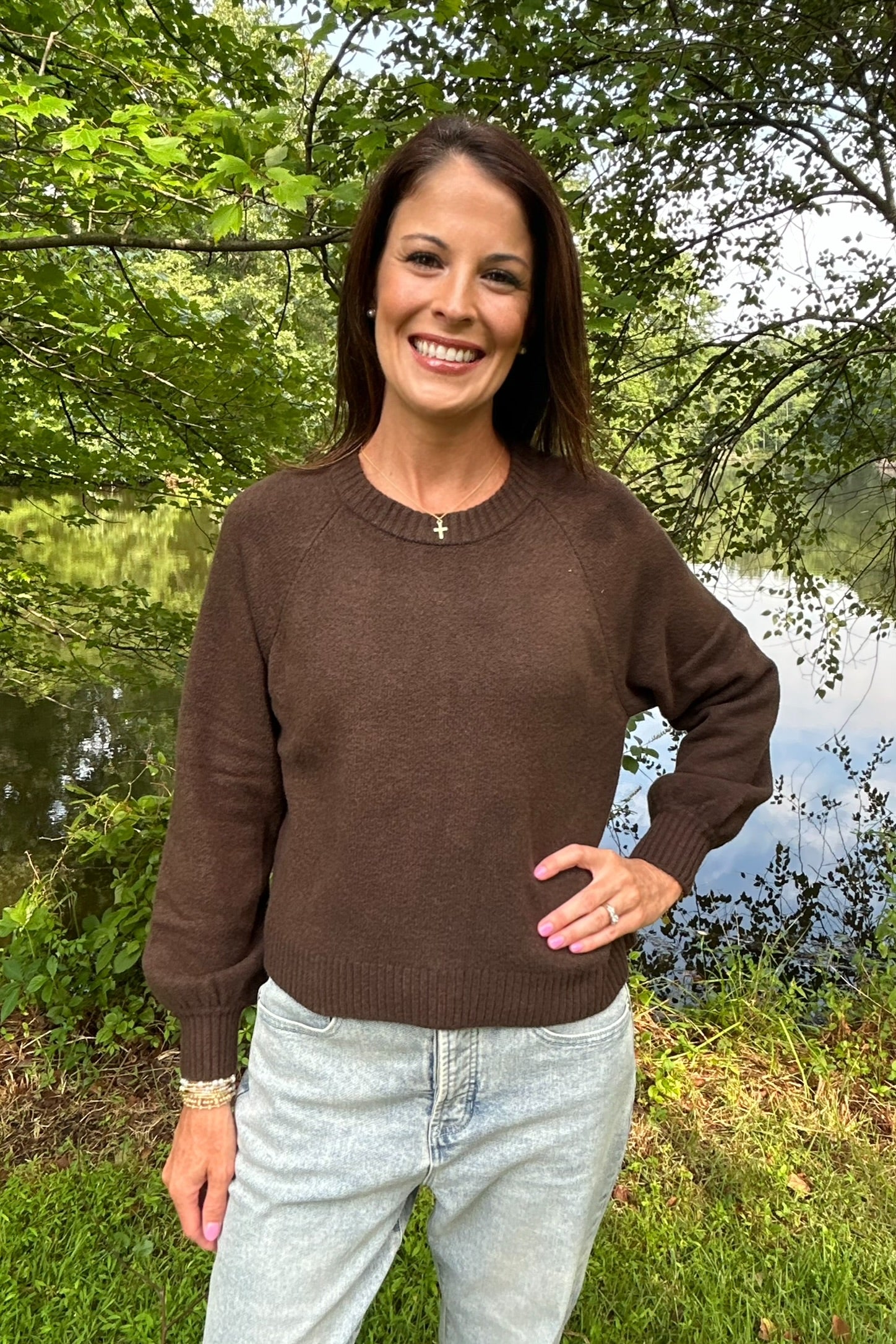 A woman smiling outdoors near a lake, wearing a brown long-sleeve crewneck sweater and light-wash jeans.