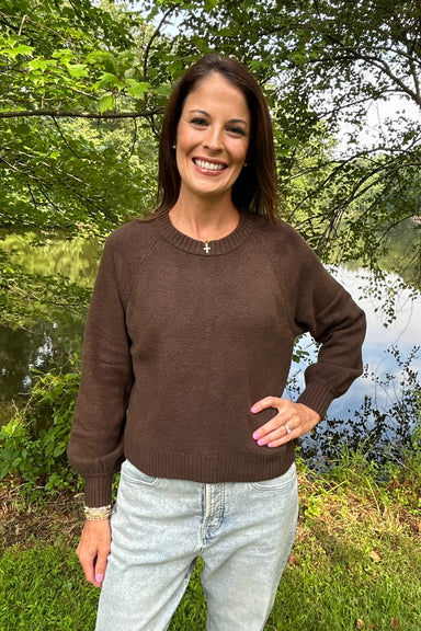 A woman smiling outdoors near a lake, wearing a brown long-sleeve crewneck sweater and light-wash jeans.