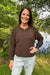 A woman smiling outdoors near a lake, wearing a brown long-sleeve crewneck sweater and light-wash jeans.