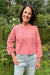 A woman wearing a dusty pink knit crewneck sweater and light-wash jeans, standing outdoors in front of a pond with lush green trees and foliage.
