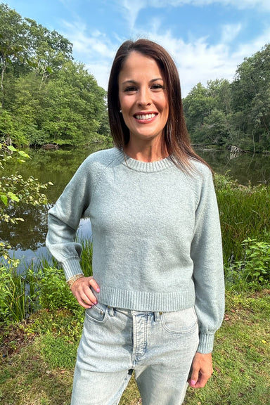 A woman smiling outdoors by a lake wearing a light sage green knit crewneck sweater and light-wash jeans.