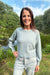 A woman smiling outdoors by a lake wearing a light sage green knit crewneck sweater and light-wash jeans.