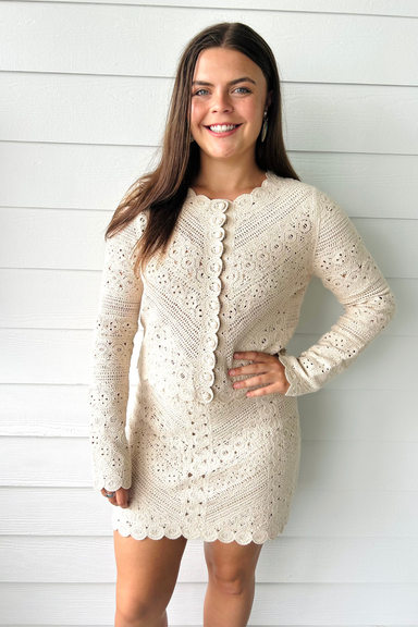 A woman wearing a cream-colored, long-sleeve crochet eyelet cardigan dress with a scalloped hem and button-front closure, standing against a white shiplap wall.